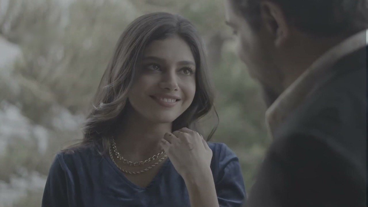 A flat, ungraded drama frame of a smiling woman in blue velvet talking to a man